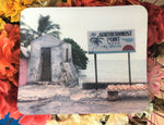 Mouse Pad showing a mid 20th century picture of the Southernmost Point: a site with a very basic rectangular signage "The Southernmost Point in U.S.A", "Key West FLA" with drawings of a palm tree, sea horizon and sunset. In front of the sign, conch shells are displayed on a bench.