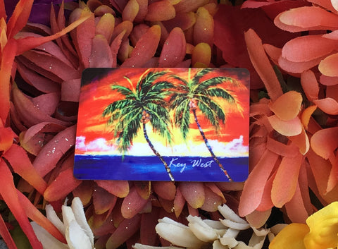 Magnet picture showing a painting of palm trees in front of a sunset waterview.