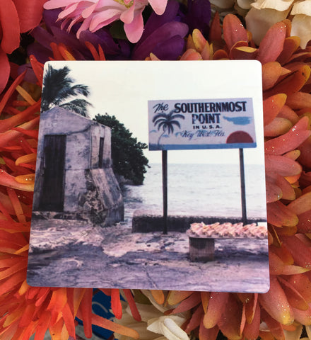 Sandstone Coaster showing a mid 20th century picture of the Southernmost Point: a site with a very basic rectangular signage "The Southernmost Point in U.S.A", "Key West FLA" with drawings of a palm tree, sea horizon and sunset. In front of the sign, conch shells are displayed on a bench.