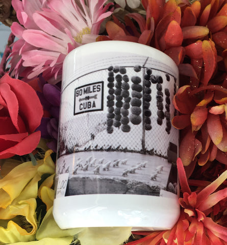 Front view mug showing a mid 20th century black and white picture of a "90 MILES TO CUBA" sign with an arrow pointing out to the sea and conch shells lined up on the cement, for sale at only $1 each! 