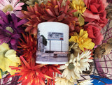 Front view mug showing a mid 20th century picture of the Southernmost Point: a site with a very basic rectangular signage "The Southernmost Point in U.S.A", "Key West FLA" with drawings of a palm tree, sea horizon and sunset. In front of the sign, conch shells are displayed on a bench.