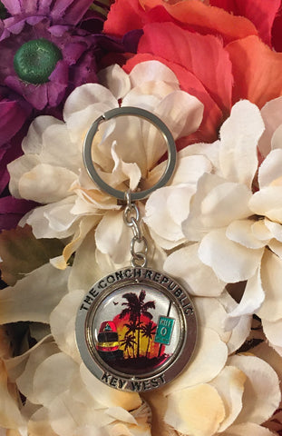 Key Chain composed of two parts: one center piece with a warm colored sunset design showing the Southernmost Point, Mile 0 and palm trees; this part rotates horizontally. And the other part around the first piece, framing it and engraved with "The Conch Republic" and "Key West".