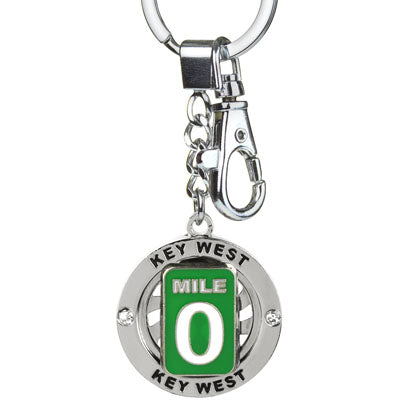 Key Chain showing a circular piece where "Key West" is engraved twice (top and bottom) and two shinny crystals are set; this circular piece is around a spinning piece in the shape and design of the Mile 0. This key chain also shows a hook (for example to attach the key chain onto a handbag).