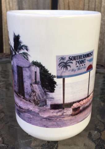 Front view mug showing a mid 20th century picture of the Southernmost Point: a site with a very basic rectangular signage "The Southernmost Point in U.S.A", "Key West FLA" with drawings of a palm tree, sea horizon and sunset. In front of the sign, conch shells are displayed on a bench.