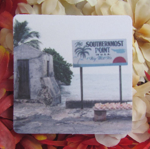 Rubber Coaster showing a mid 20th century picture of the Southernmost Point: a site with a very basic rectangular signage "The Southernmost Point in U.S.A", "Key West FLA" with drawings of a palm tree, sea horizon and sunset. In front of the sign, conch shells are displayed on a bench.