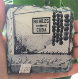 Slate Coaster showing a mid 20th century black and white picture of a "90 MILES TO CUBA" sign with an arrow pointing out to the sea and conch shells lined up on the cement, for sale at only $1 each! 