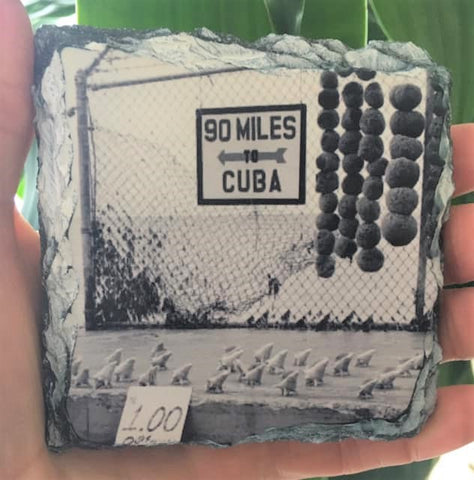 Slate Coaster showing a mid 20th century black and white picture of a "90 MILES TO CUBA" sign with an arrow pointing out to the sea and conch shells lined up on the cement, for sale at only $1 each! 